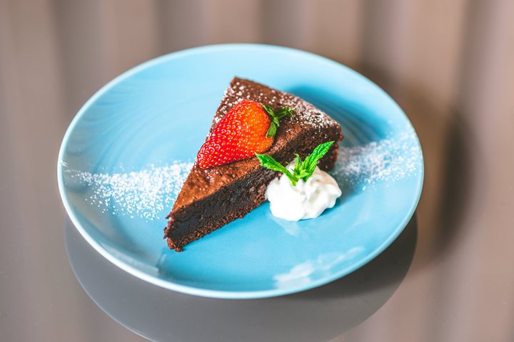 Kawałek brownie z truskawką, bitą śmietaną i cukrem pudrem na niebieskim talerzyku