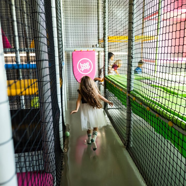 Dziewczynka w białej spódniczce biegnie korytarzem konstrukcji obok trampolin